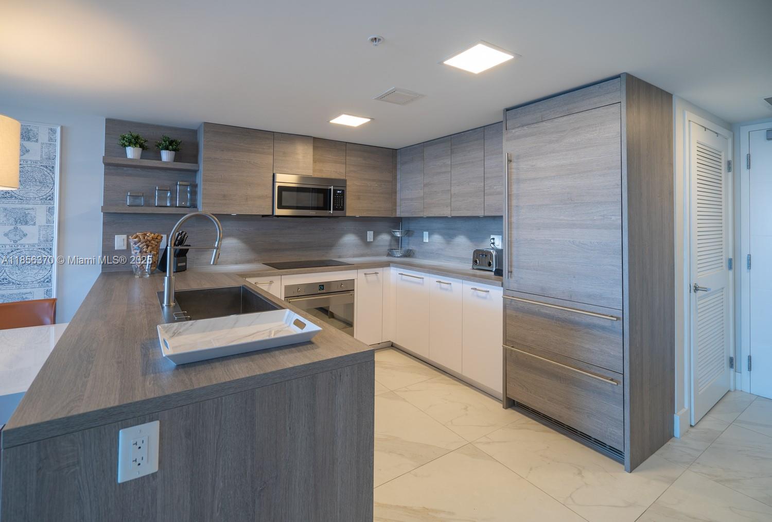 A modern kitchen features wood-grain cabinetry, white lower cabinets, stainless steel appliances, and marble flooring — SUNNY ISLES BEACH, FL