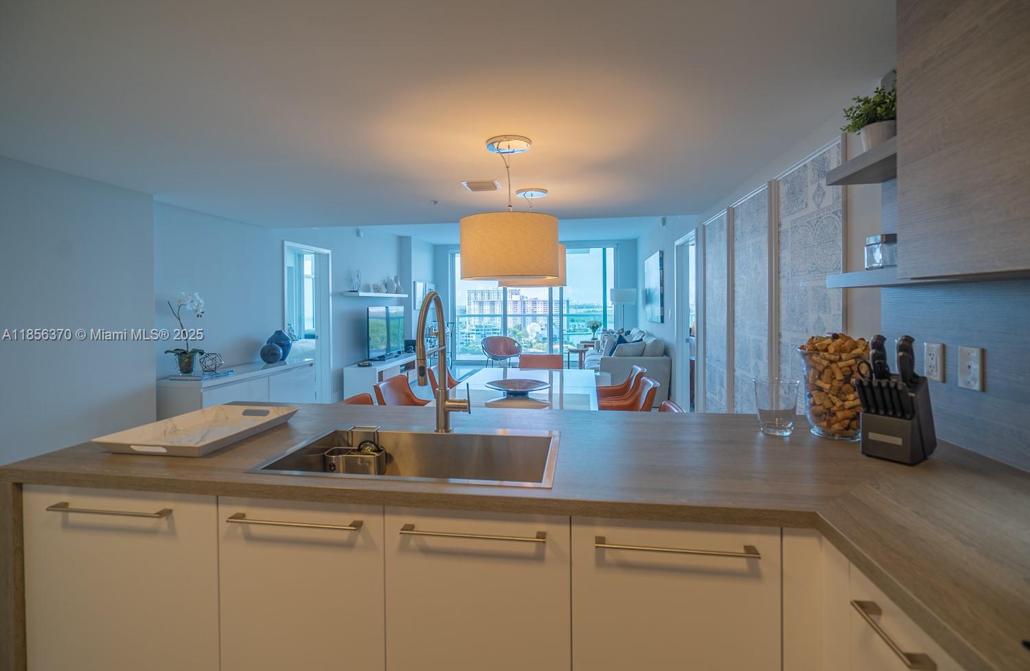 A modern kitchen with white cabinets and wood-tone countertops overlooks an open-concept living area — SUNNY ISLES BEACH, FL