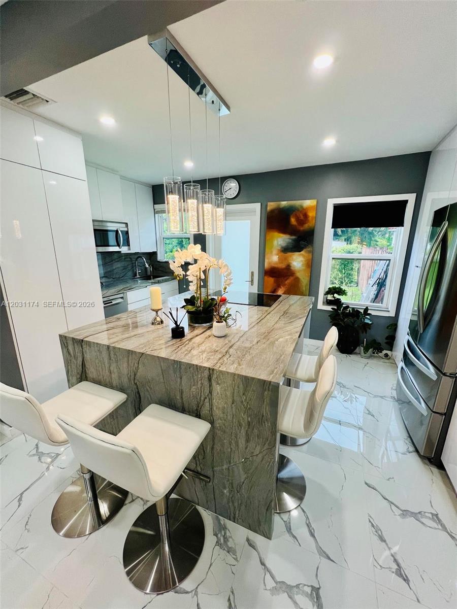 A modern kitchen features a large stone-topped island with white bar stools and marble flooring — 920 NE 108th St # 0, Biscayne Park FL 33161, BISCAYNE PARK, FL