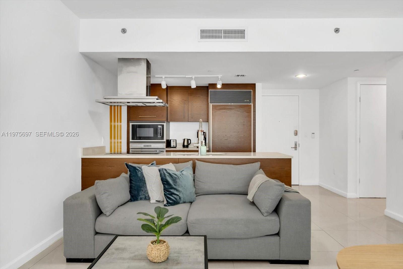 A gray sofa sits in front of a kitchen with wood cabinets and stainless appliances — 485 Brickell Ave # 1807, Miami FL 33131, MIAMI, FL