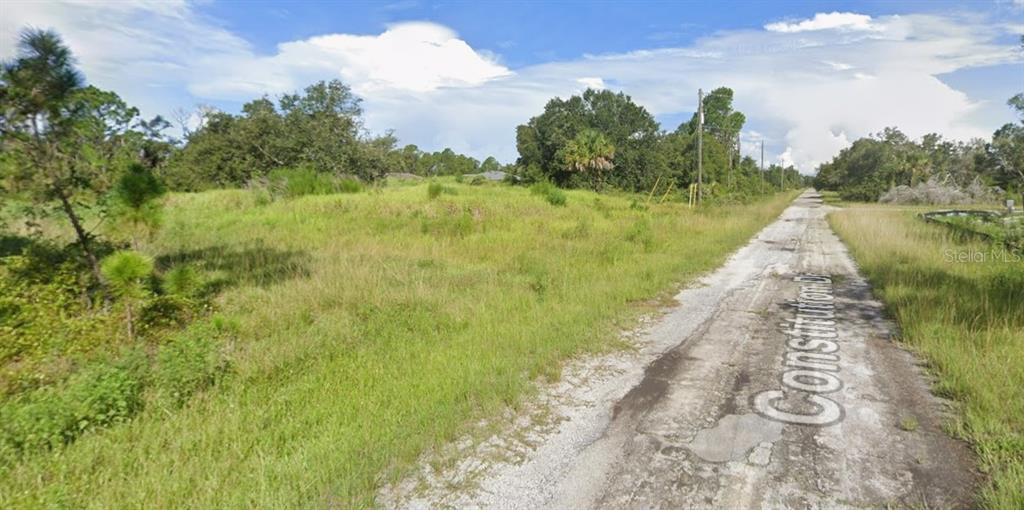 A gravel road runs alongside a grassy, undeveloped lot with scattered trees and shrubs — Lot 69 CONSTITUTION DRIVE, NORTH PORT, FL