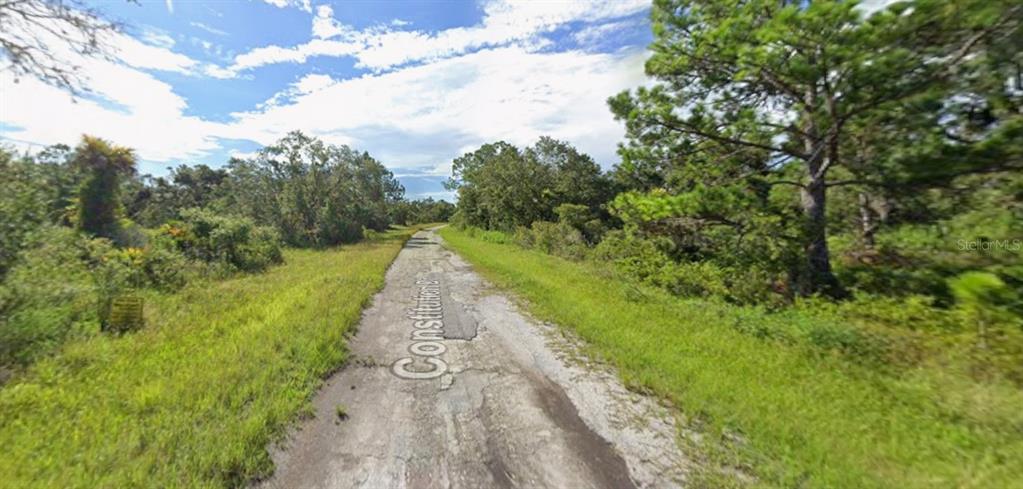 A narrow, weathered asphalt road stretches through a rural area surrounded by trees and vegetation — Lot 69 CONSTITUTION DRIVE, NORTH PORT, FL