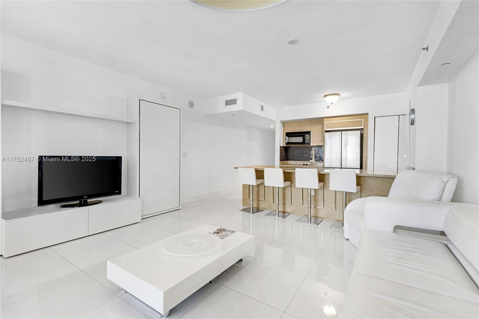 A modern living room features white tile flooring, a television, and an open kitchen area — 1830 S Ocean Dr # 2609, Hallandale Beach FL 33009, HALLANDALE BEACH, FL