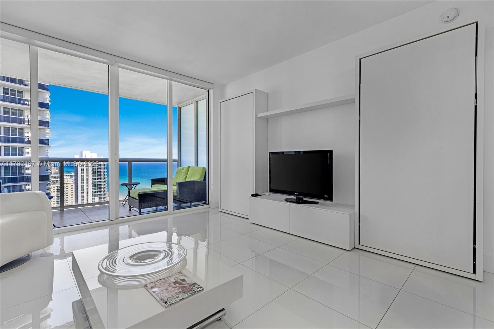 A modern living room features white tile floors, a wall-mounted television, and a balcony view — 1830 S Ocean Dr # 2609, Hallandale Beach FL 33009, HALLANDALE BEACH, FL