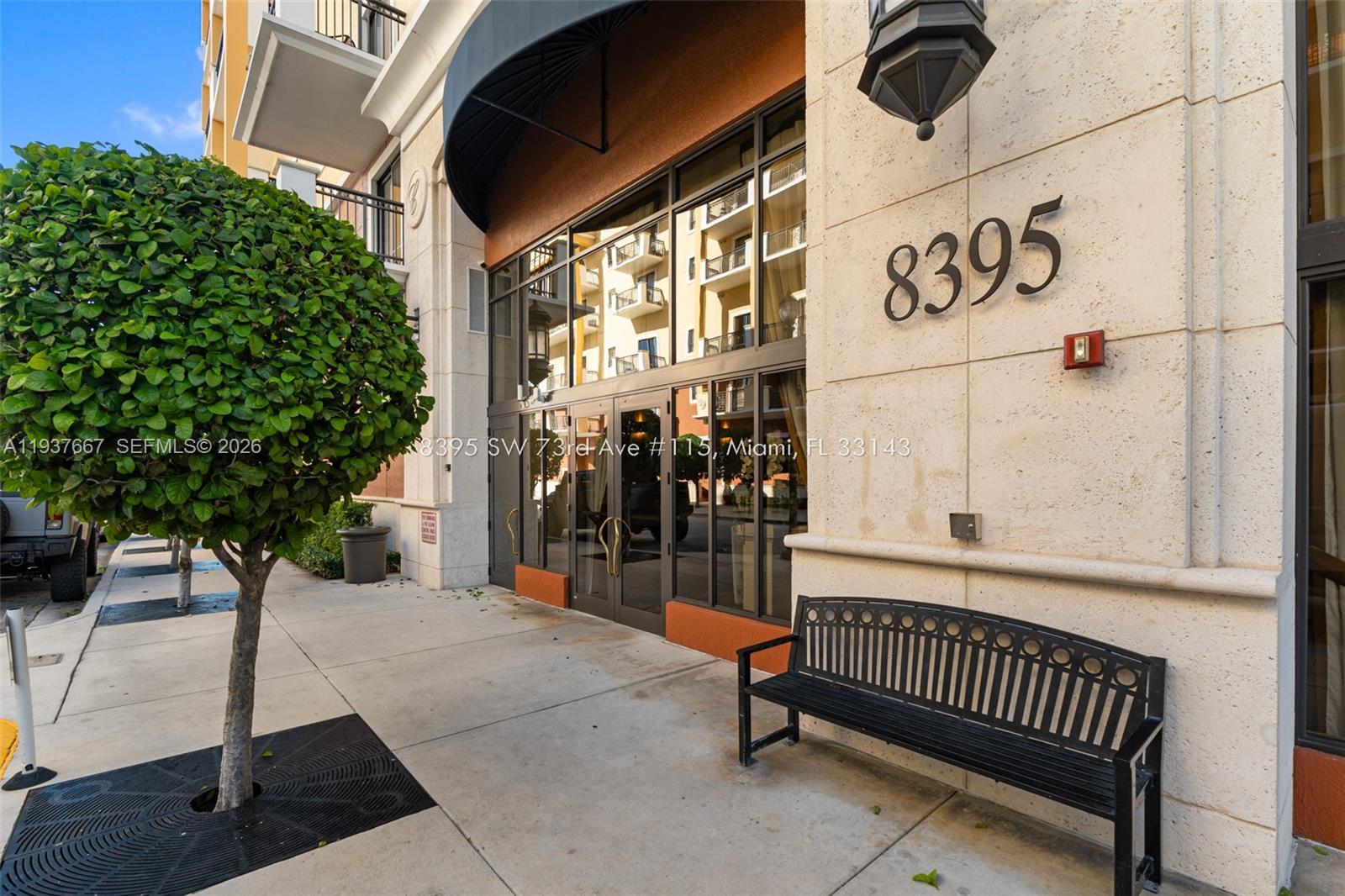 The exterior entrance of a building features a glass door, a black bench, and landscaping — 8395 SW 73rd Ave # 115, Miami FL 33143, MIAMI, FL