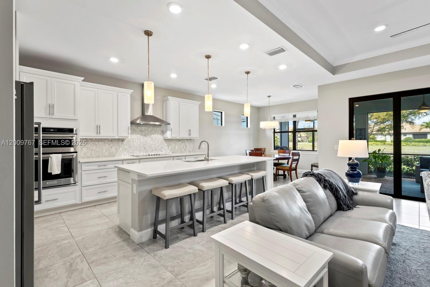 A modern kitchen features white cabinetry, a large island with bar seating, and an adjacent living area — 6359 Achievement, Ave Maria FL 34142, AVE MARIA, FL