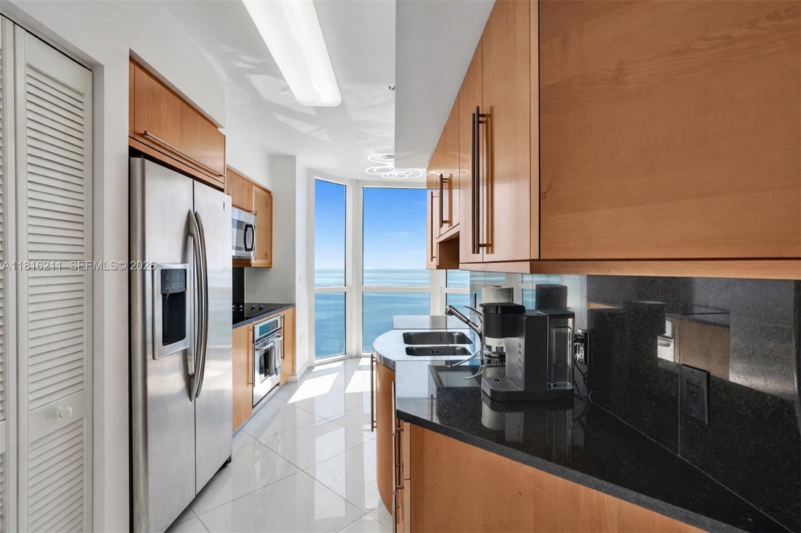 A modern kitchen features light wood cabinets, black granite countertops, and floor-to-ceiling ocean views — 6365 Collins Ave # 2403, Miami Beach FL 33141, MIAMI BEACH, FL