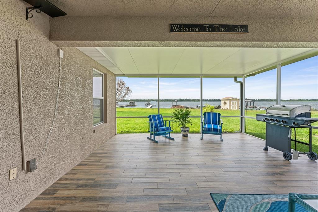 A covered patio with wood-look tile flooring overlooks a lake with a grill and chairs — 4111 ISLAND LAKES DRIVE, WINTER HAVEN, FL