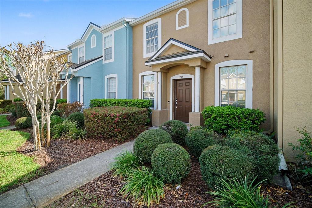A tan townhouse exterior with a brown front door, a small porch, and landscaped bushes — 1246 CHARLESWORTH DRIVE, WESLEY CHAPEL, FL