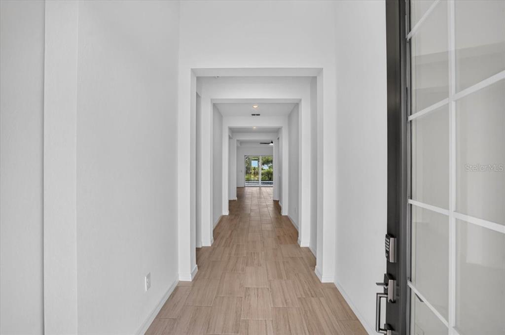 A long, white-walled hallway with light wood-look tile flooring leads toward a glass-paned exterior door — 19219 SCALLOP LOOP, LAKEWOOD RANCH, FL