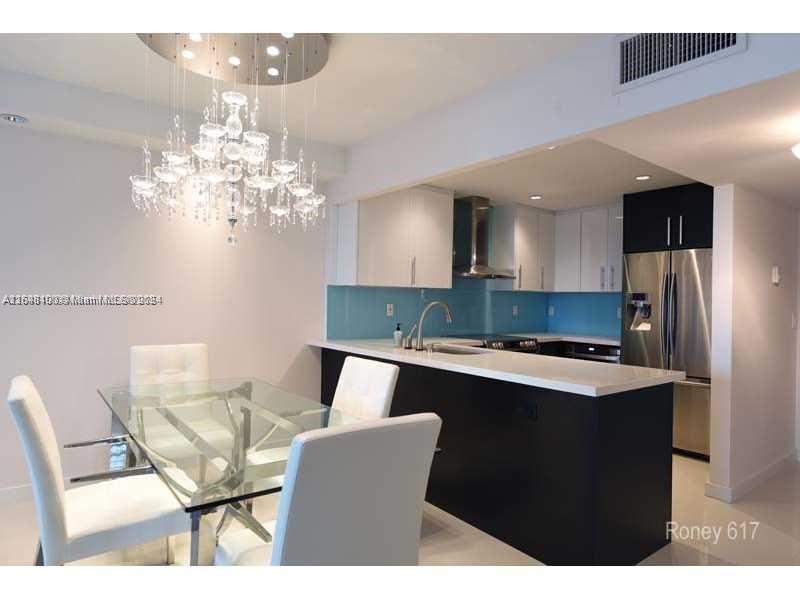 A dining area with a glass table and chandelier sits beside a modern kitchen island — 2301 Collins Ave # 617, Miami Beach FL 33139, MIAMI BEACH, FL