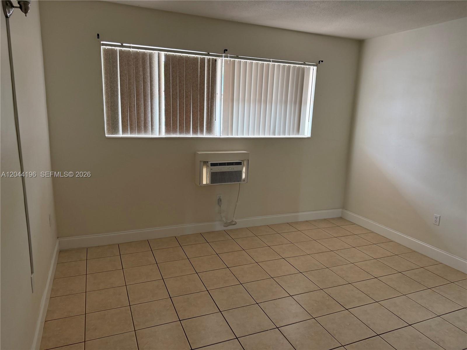 An empty room features beige tile flooring, a window with vertical blinds, and a wall-mounted air conditioner — 19001 NE 2nd Ave # 1404, Miami FL 33179, MIAMI, FL
