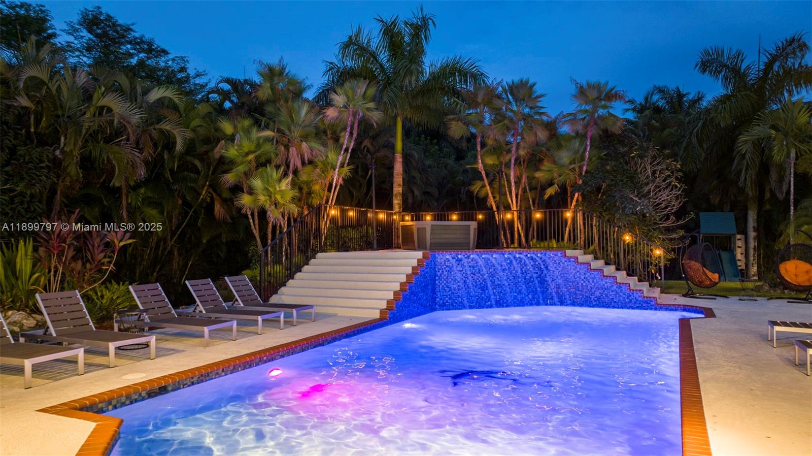 A swimming pool with a blue tiled waterfall feature and lounge chairs at dusk — 3152 SW 147th Ave, Davie FL 33330, DAVIE, FL
