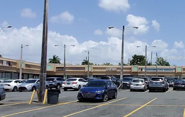 A parking lot sits in front of a strip mall with various storefronts under a blue sky — Forest Hills PR-167 INT. CLL-15, BAYAMON, PR
