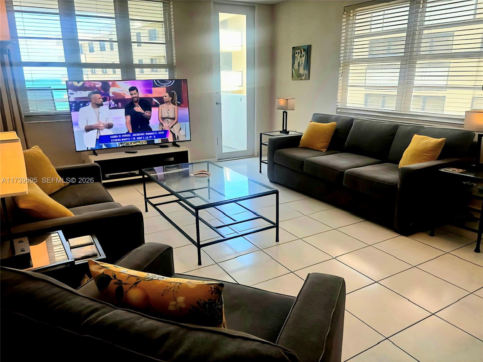 A living room features dark gray sofas, a glass coffee table, and large windows with blinds — 100 Lincoln Rd # 821, Miami Beach FL 33139, MIAMI BEACH, FL