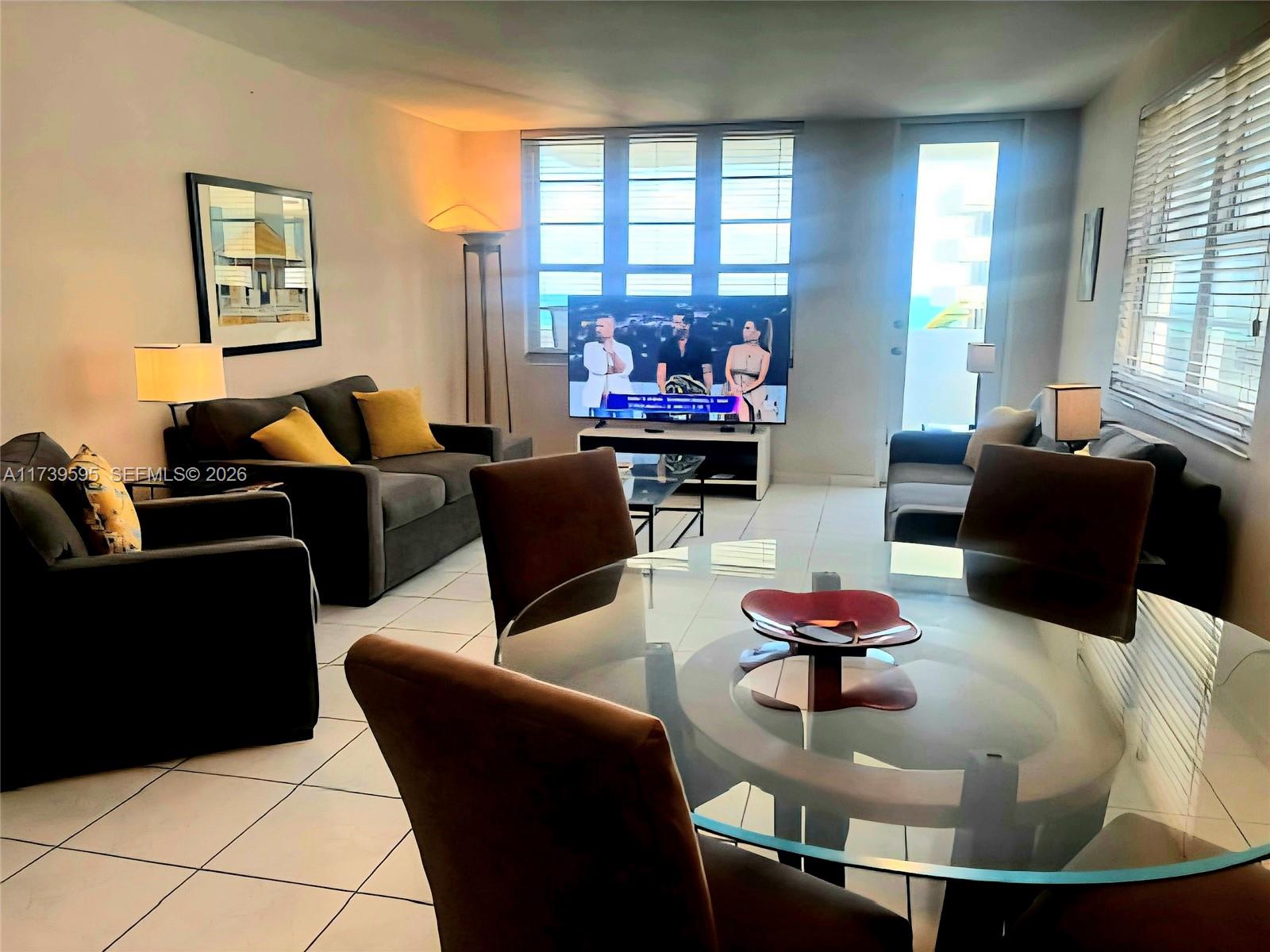 A living and dining area features a glass table, gray sofas, and a television — 100 Lincoln Rd # 821, Miami Beach FL 33139, MIAMI BEACH, FL