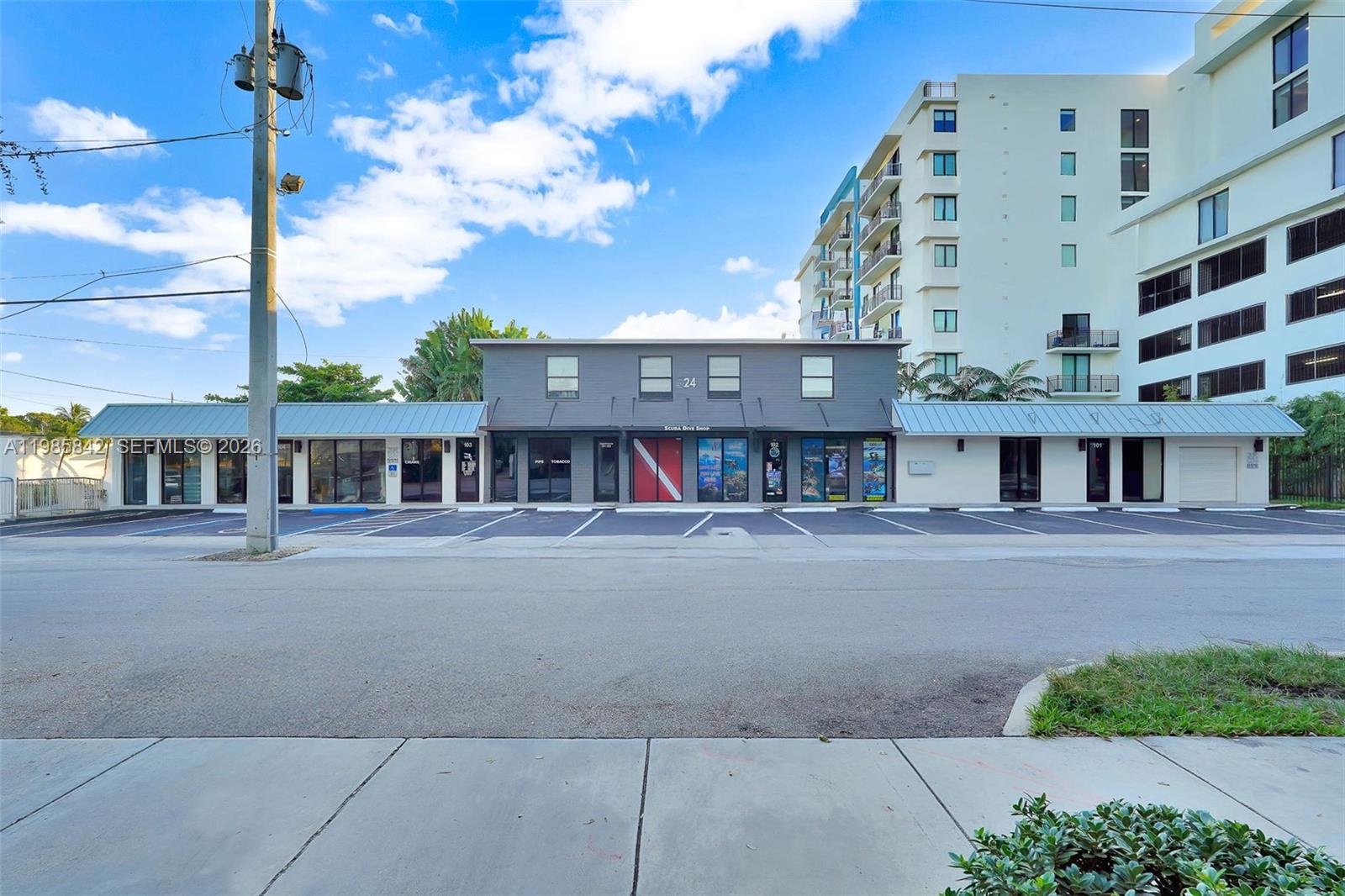 A low-rise commercial building with storefronts and a parking lot sits beside a tall apartment — 24 NE 1st Ave, Dania Beach FL 33004, DANIA BEACH, FL
