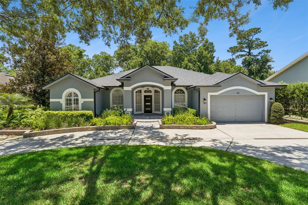 A gray single-story house with a two-car garage and a curved driveway under large trees — 148 WISTERIA DRIVE, LONGWOOD, FL