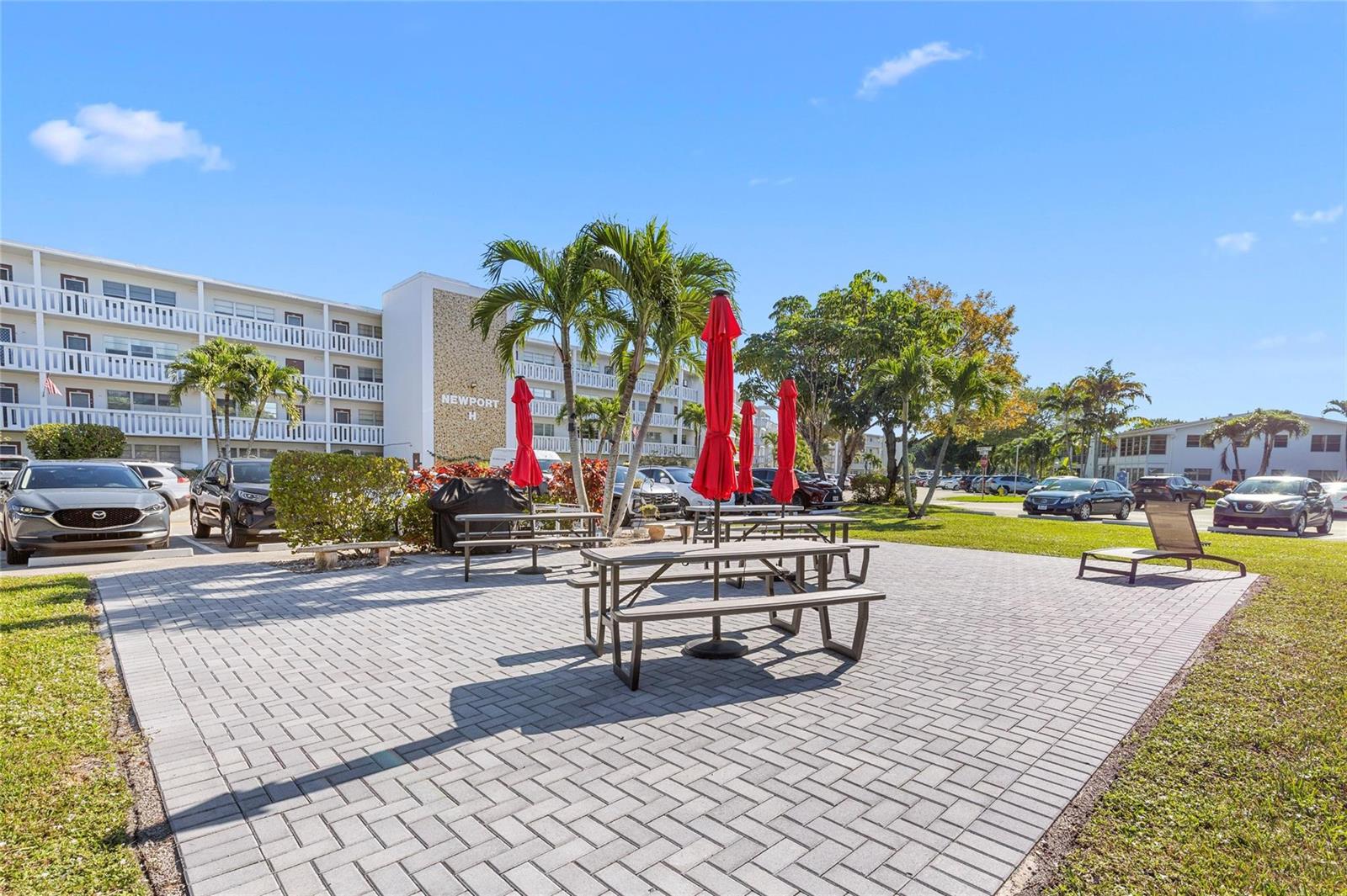 A paved patio area features picnic tables with red umbrellas in front of apartment buildings — 3028 Newport H, Deerfield Beach FL 33442, DEERFIELD BEACH, FL