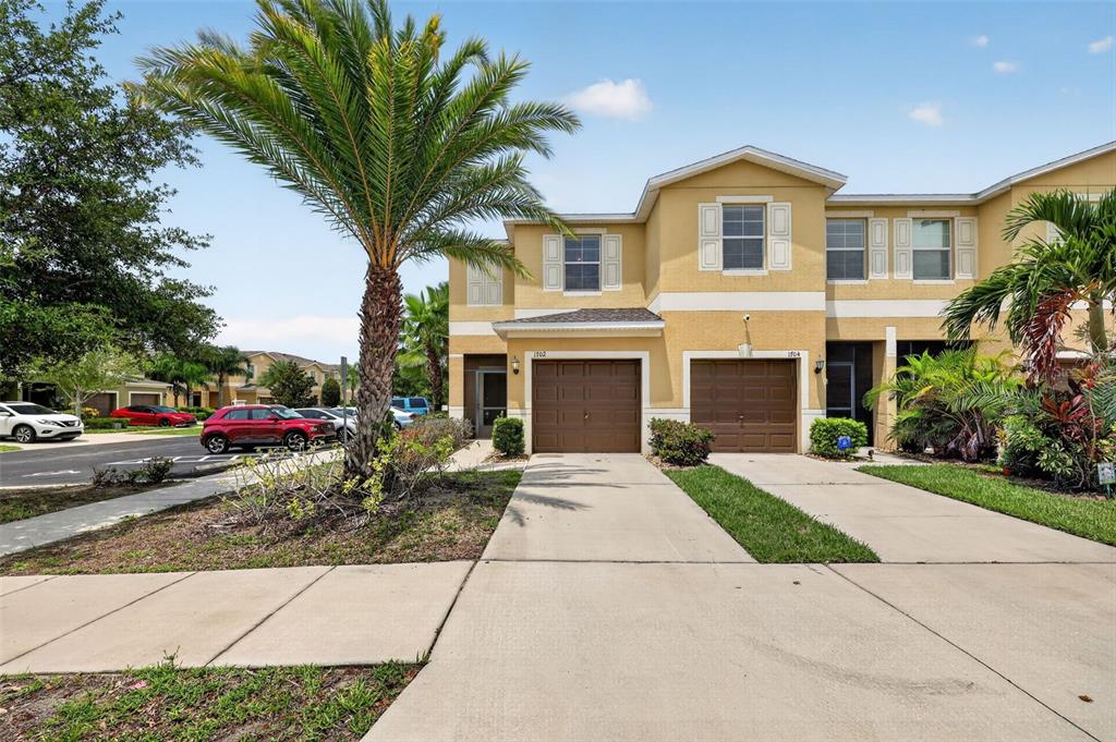 A two-story townhouse exterior features tan stucco walls, brown garage doors, and a palm tree — 1702 IVORY GOOSE PLACE, RUSKIN, FL