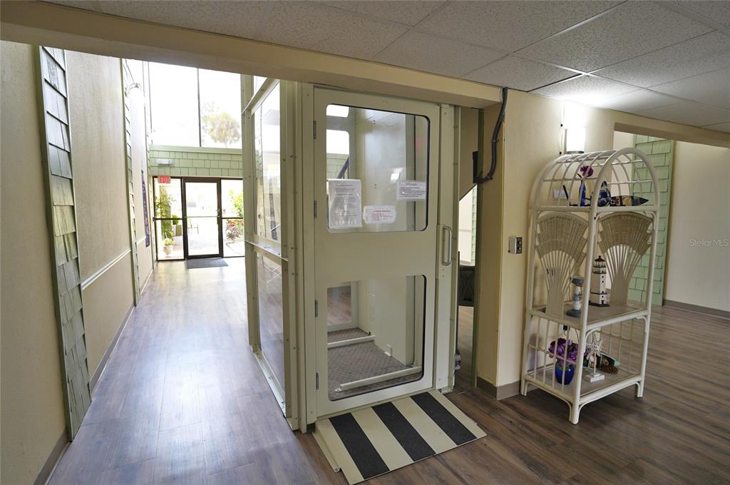 An indoor wheelchair lift stands in a hallway with wood-look flooring and a wicker shelf — 21267 GERTRUDE AVENUE UNIT 210, PORT CHARLOTTE, FL