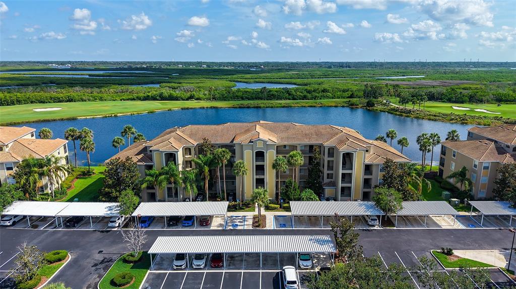 An aerial view shows a multi-story apartment complex with covered parking spaces beside a lake — 7803 GRAND ESTUARY TRAIL UNIT 307, BRADENTON, FL