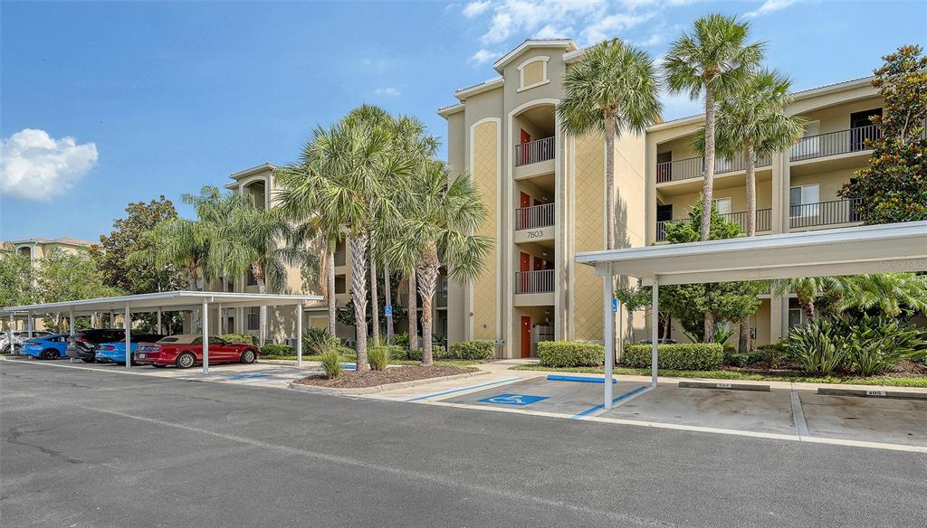 A multi-story apartment building with covered parking spaces and palm trees in the foreground — 7803 GRAND ESTUARY TRAIL UNIT 307, BRADENTON, FL