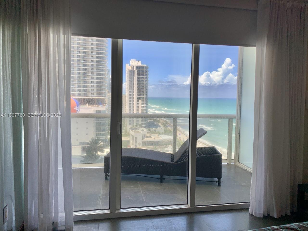 A balcony with a lounge chair overlooks the ocean and high-rise buildings through glass doors — 1830 S Ocean Dr # 1610, Hallandale Beach FL 33009, HALLANDALE BEACH, FL