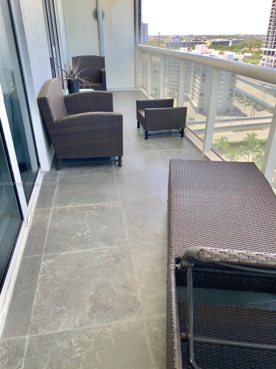 A balcony features gray tile flooring, wicker furniture, and a glass railing overlooking a city — 1830 S Ocean Dr # 1610, Hallandale Beach FL 33009, HALLANDALE BEACH, FL