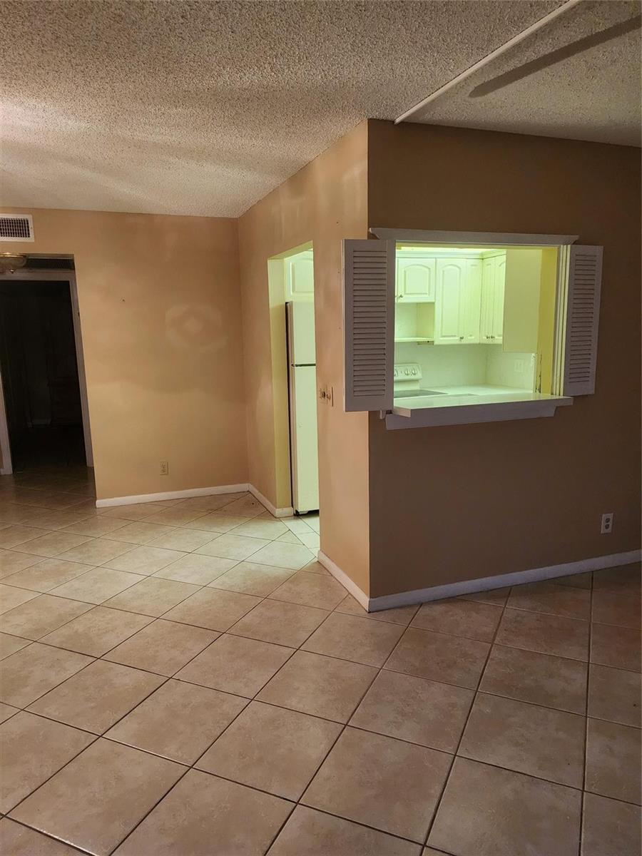 A room with tan walls and tile flooring features a kitchen pass-through window with shutters — 104 Westbury E, Deerfield Beach FL 33442, DEERFIELD BEACH, FL
