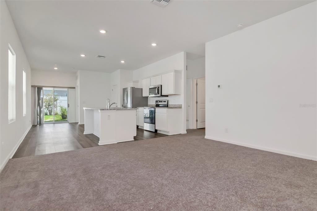 A living room with gray carpet transitions into a kitchen featuring white cabinets and an island — 35575 BUTTONWEED TRL, ZEPHYRHILLS, FL