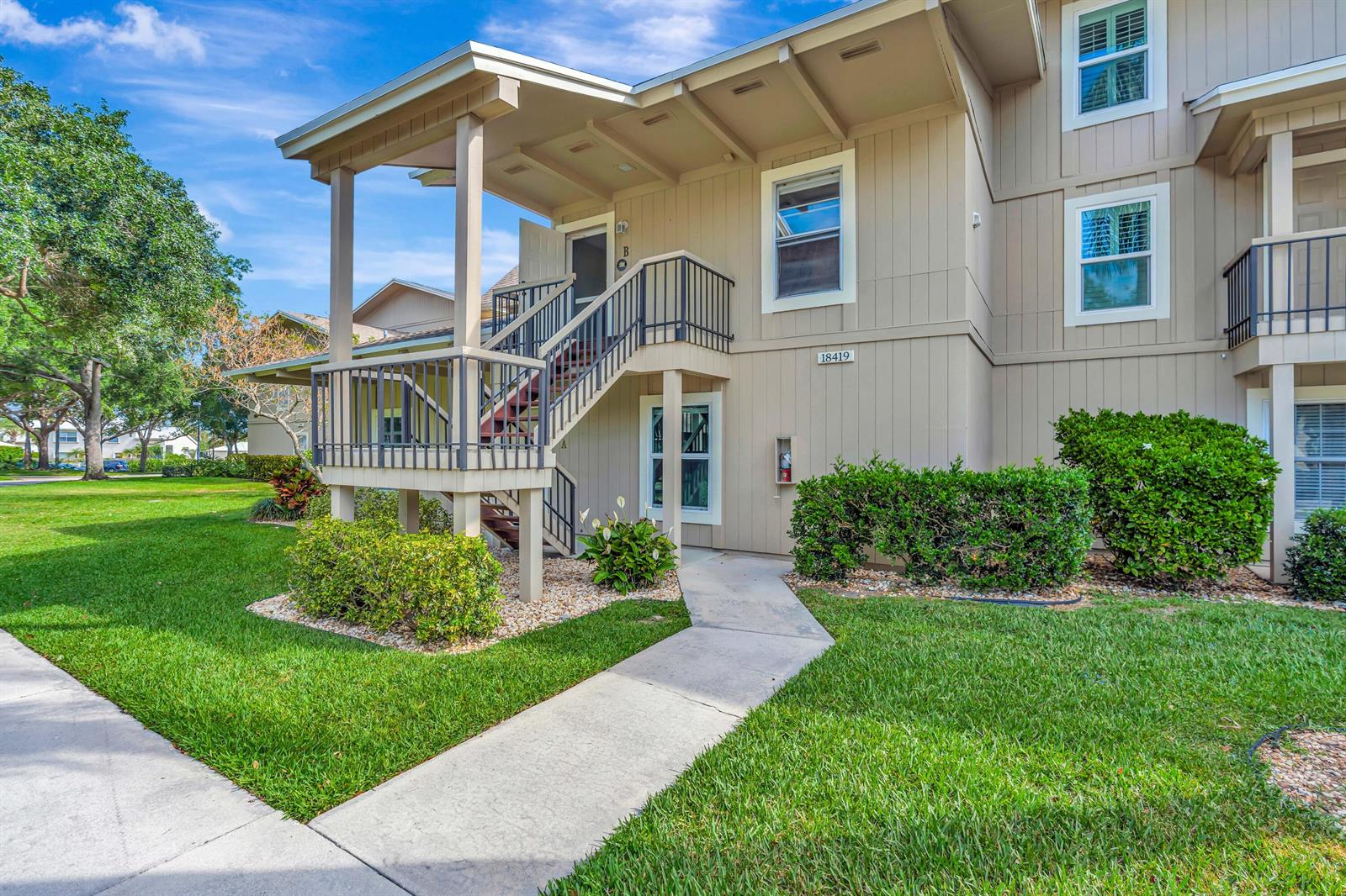 An exterior view of a two-story tan apartment building with a staircase leading to unit B — 18419 SE Wood Haven Lane # B, Tequesta FL 33469, TEQUESTA, FL