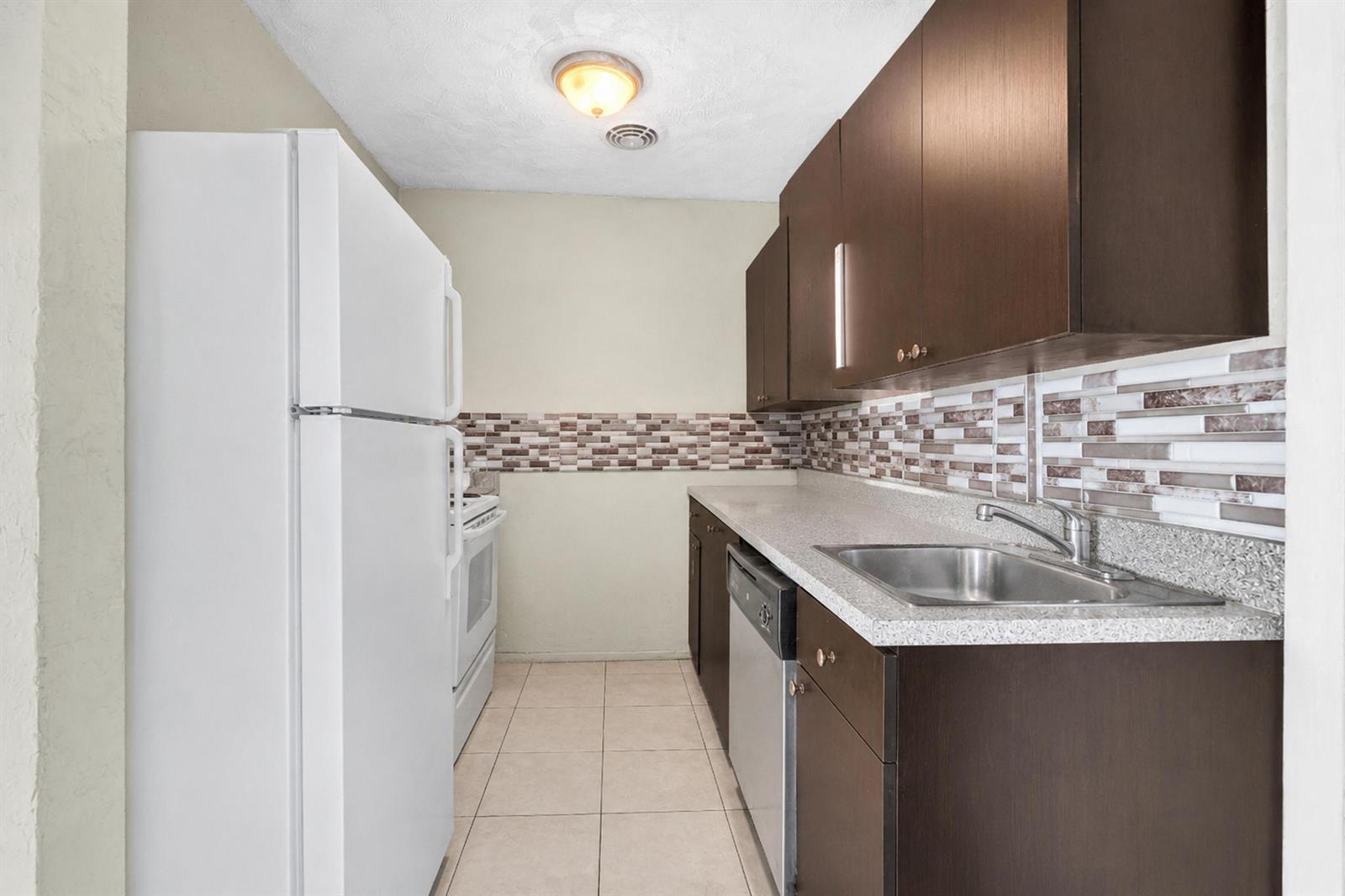 A galley kitchen features dark wood cabinets, white appliances, a tiled backsplash, and light flooring — 2743 NW 2nd Street, Boynton Beach FL 33435, BOYNTON BEACH, FL