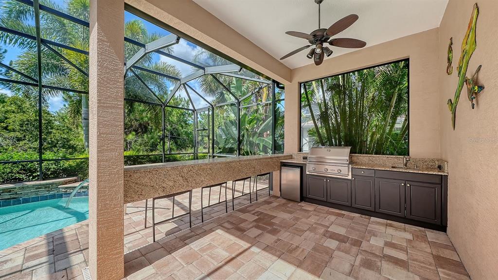 An outdoor kitchen with a grill, sink, and bar seating overlooks a screened-in swimming pool — 7640 HADDINGTON COVE, LAKEWOOD RANCH, FL