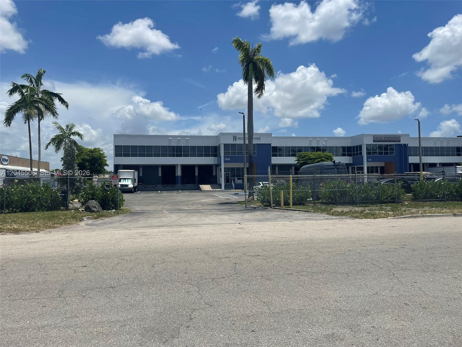A two-story commercial warehouse building features loading docks, a chain-link fence, and palm trees — FL