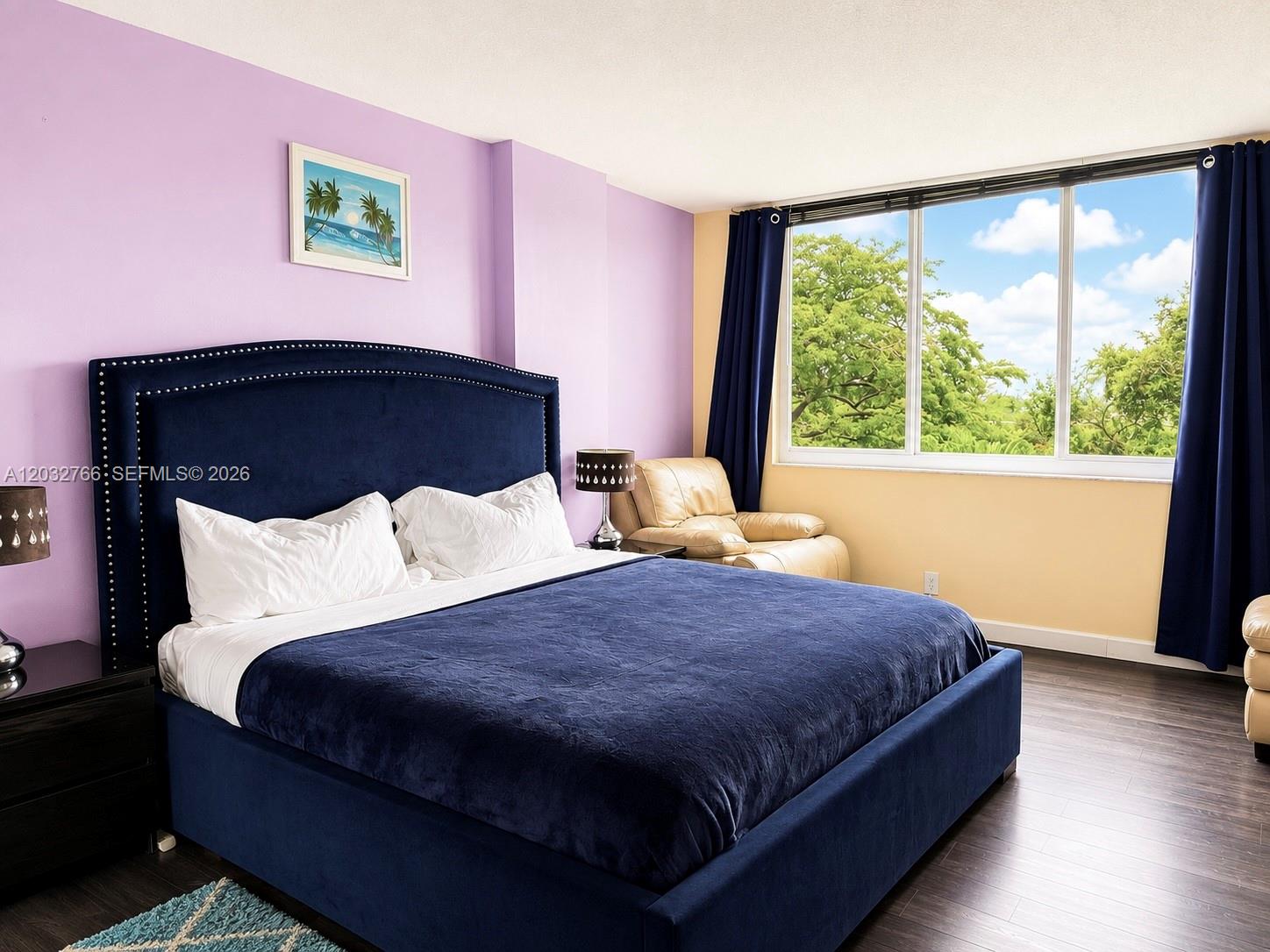 A bedroom features a blue upholstered bed, lavender walls, and a large window with trees — 4200 Hillcrest Dr # 403, Hollywood FL 33021, HOLLYWOOD, FL