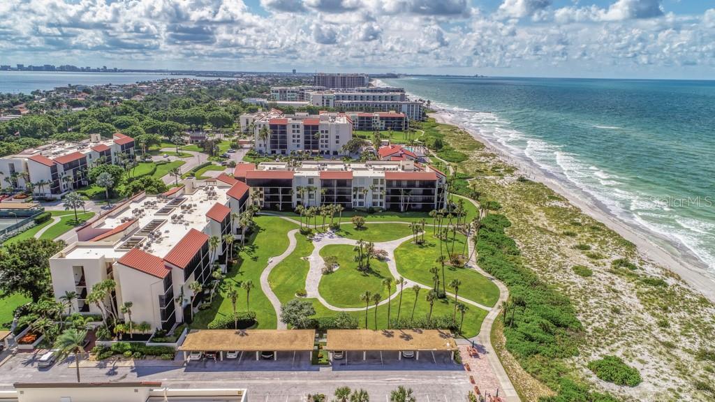 An aerial view shows beachfront condominium buildings with red roofs, landscaped grounds, and parking structures — 1975 GULF OF MEXICO DRIVE UNIT G4-401, LONGBOAT KEY, FL
