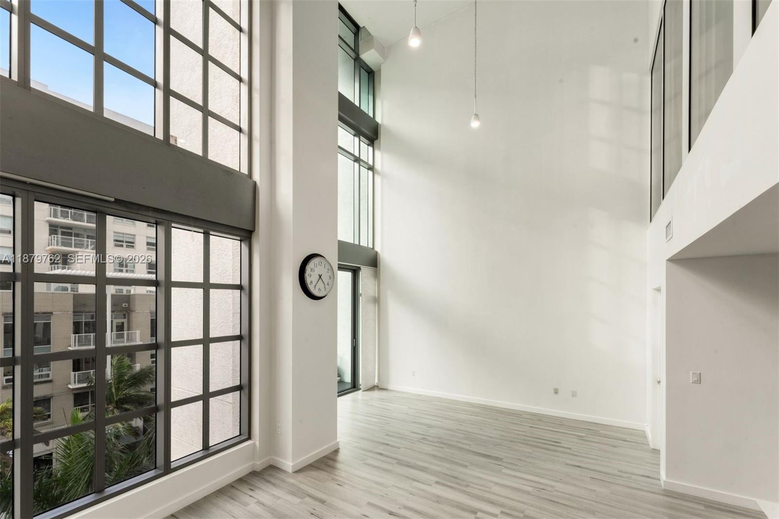 A bright, double-height living room features floor-to-ceiling windows, light wood flooring, and a wall clock — 3301 NE 1st Ave # M0501, Miami FL 33137, MIAMI, FL