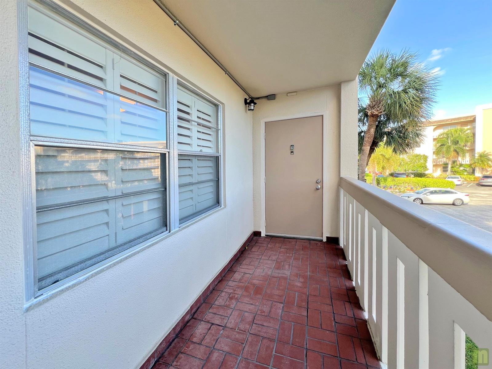 A covered balcony features red brick flooring, a beige door, and white railings — 1901 Bermuda Circle # M2, Coconut Creek FL 33066, COCONUT CREEK, FL
