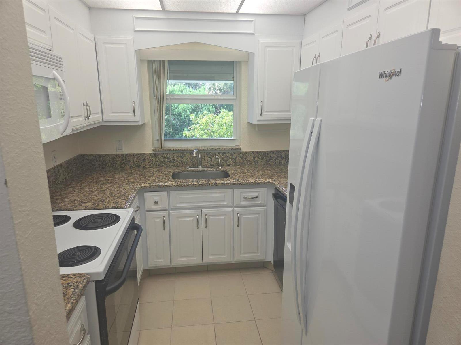 A kitchen features white cabinets, granite countertops, a white refrigerator, and a window above the sink — 1340 NE 28th Avenue # 228, Pompano Beach FL 33062, POMPANO BEACH, FL