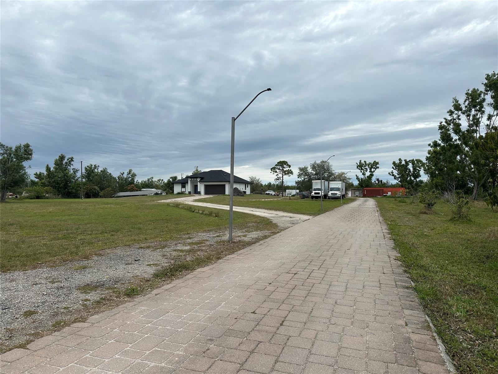 A paved driveway leads to a white house with a dark roof under a cloudy sky — 3401 Sand Road, Cape Coral FL 33993, CAPE CORAL, FL
