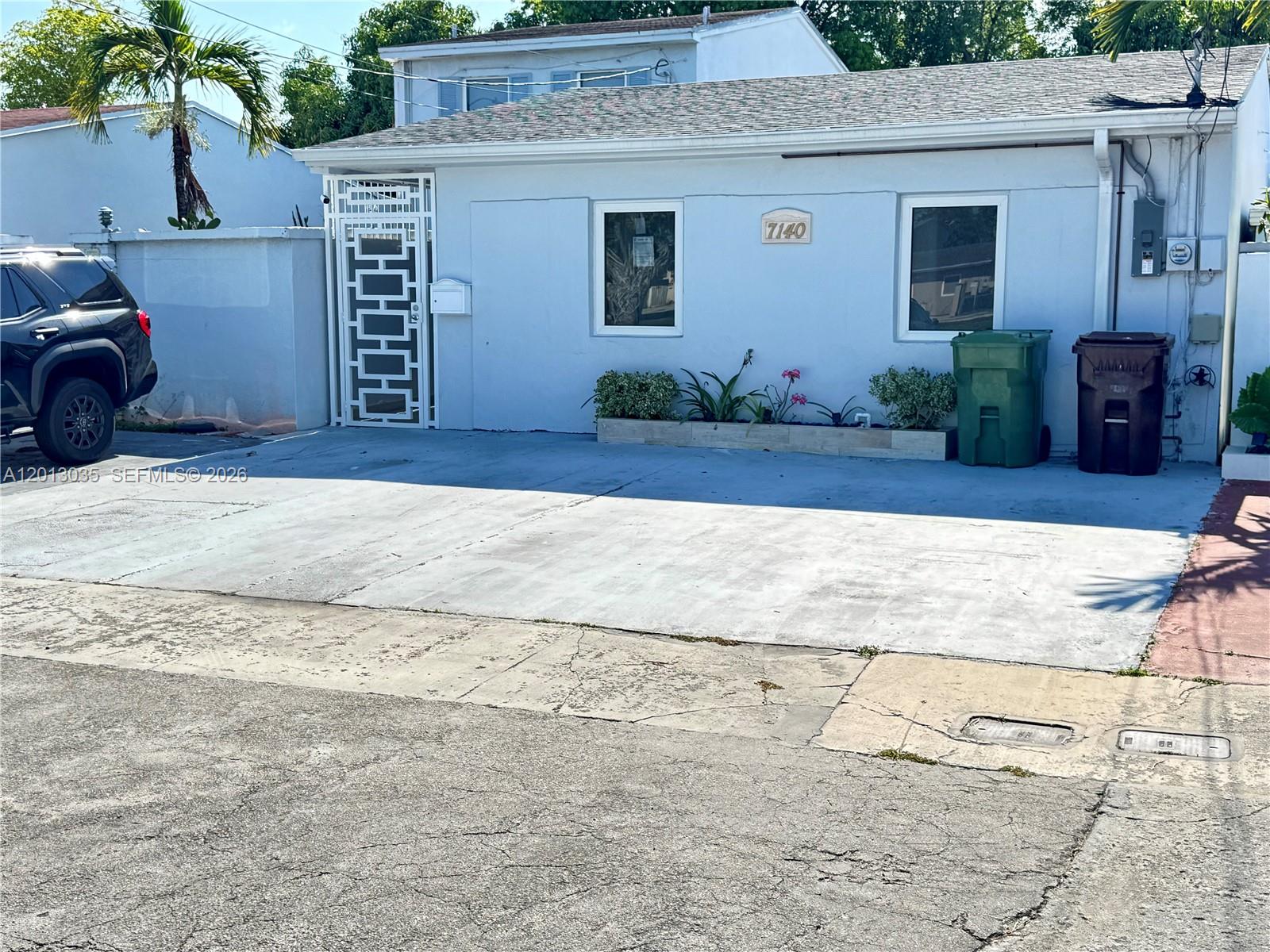 A light blue single-story house features a concrete driveway, a white gate, and two trash bins — 7140 W 15th Ct, Hialeah FL 33014, HIALEAH, FL