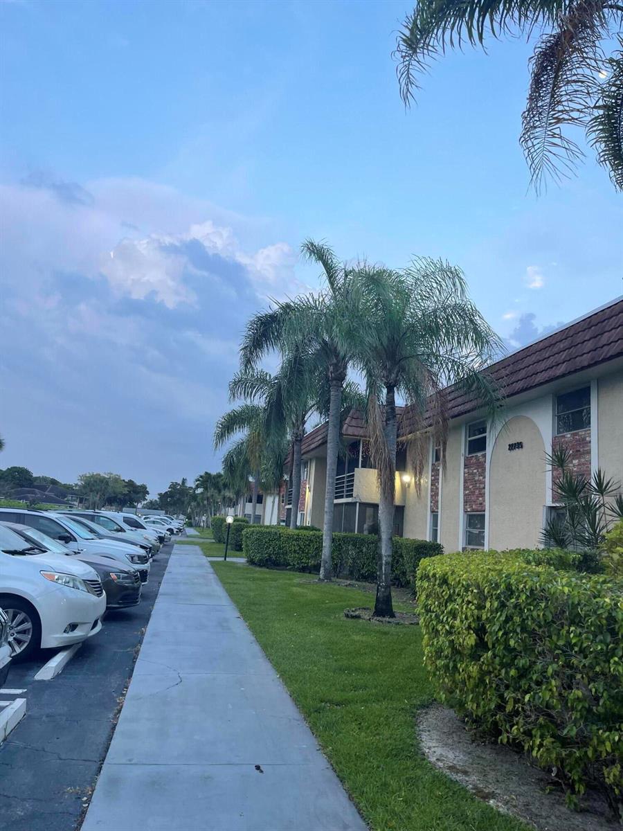 A sidewalk runs alongside a multi-story apartment building with palm trees and parked cars — 22735 SW 66th Avenue, Boca Raton FL 33428, BOCA RATON, FL