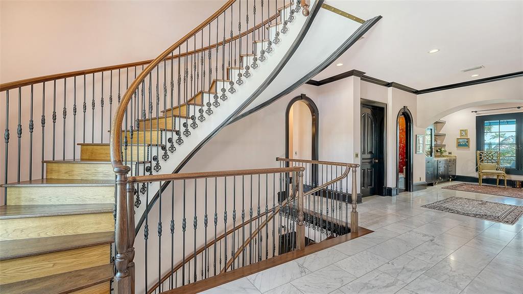 A curved wooden staircase with iron railings stands in a marble-floored foyer with arched doorways — 513 VILLA ROSA WAY, ANNA MARIA, FL