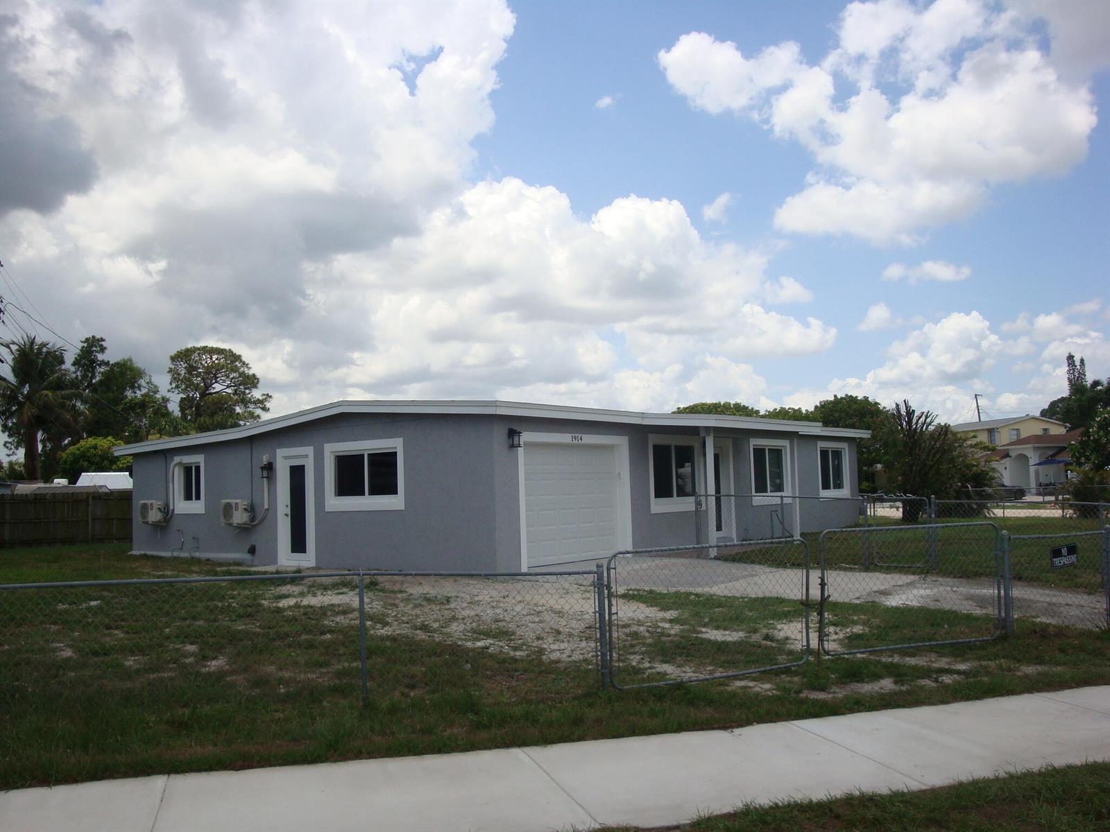 A gray, single-story house with a white garage door sits behind a chain-link fence — 1914 Kudza Road, West Palm Beach FL 33415, WEST PALM BEACH, FL