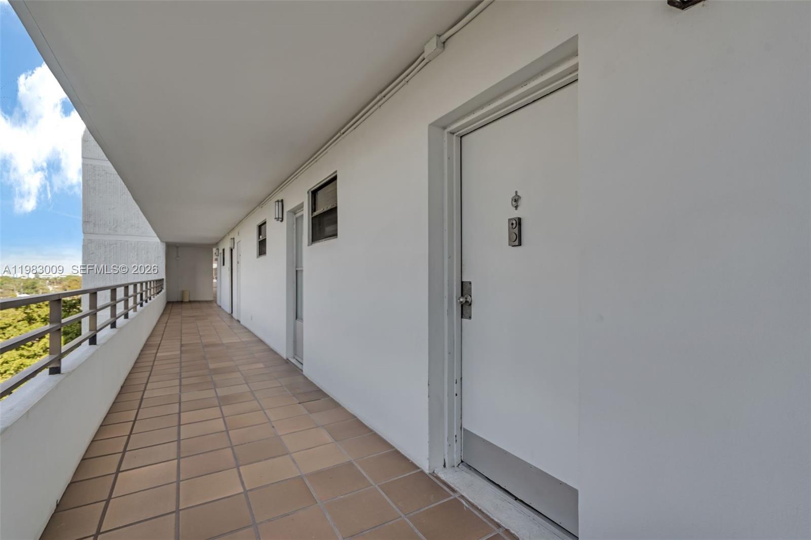 An outdoor apartment building walkway features tiled flooring, white walls, and a metal railing — 500 Three Islands Blvd # 226, Hallandale Beach FL 33009, HALLANDALE BEACH, FL
