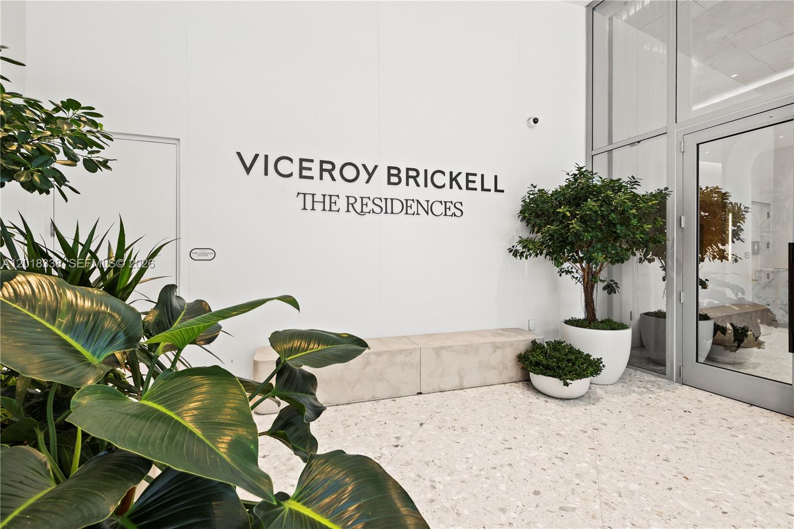 A modern building entrance features a white wall with signage, potted plants, and a bench — 77 SE 5TH # 2404, Miami FL 33131, MIAMI, FL