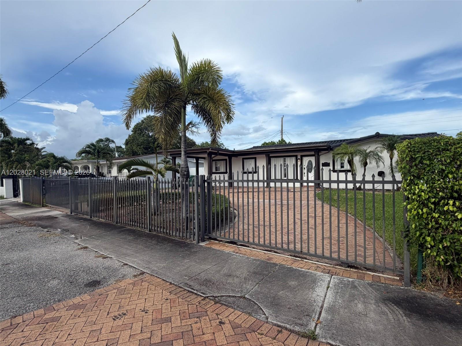 A single-story house with a brick driveway is enclosed by a tall metal fence — MIAMI GARDENS, FL