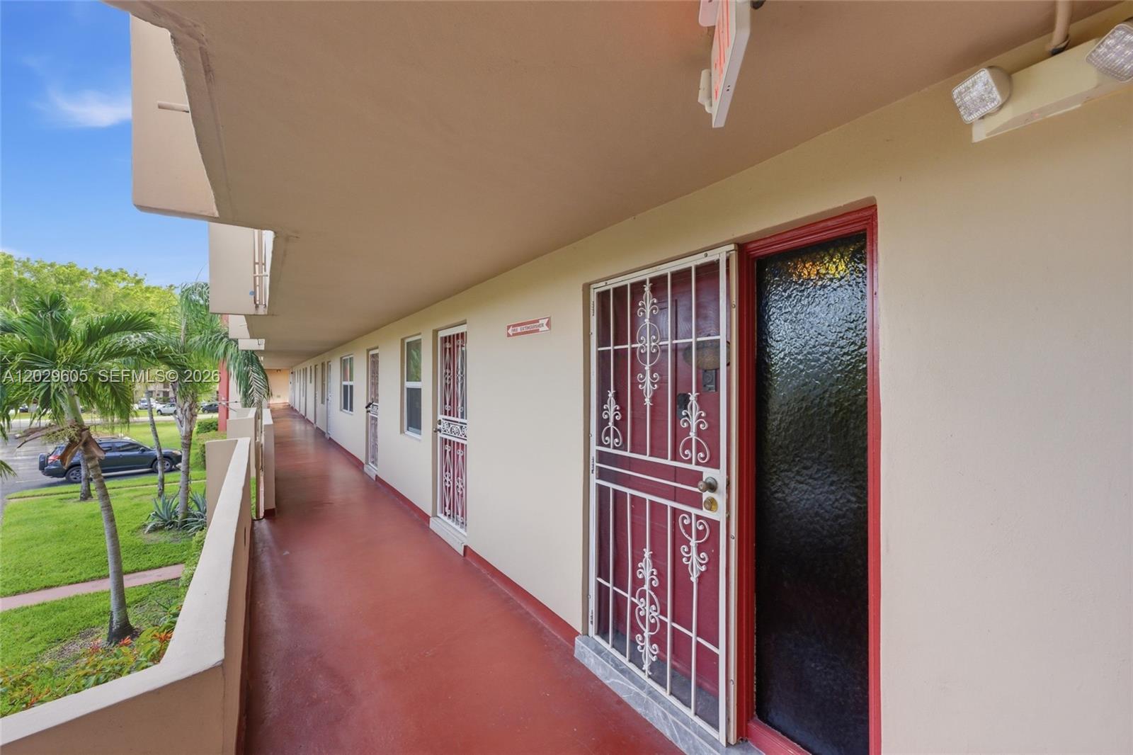 An outdoor apartment building walkway features red flooring, beige walls, and white security screen doors — 1501 NE 191st St # C217, Miami FL 33179, MIAMI, FL