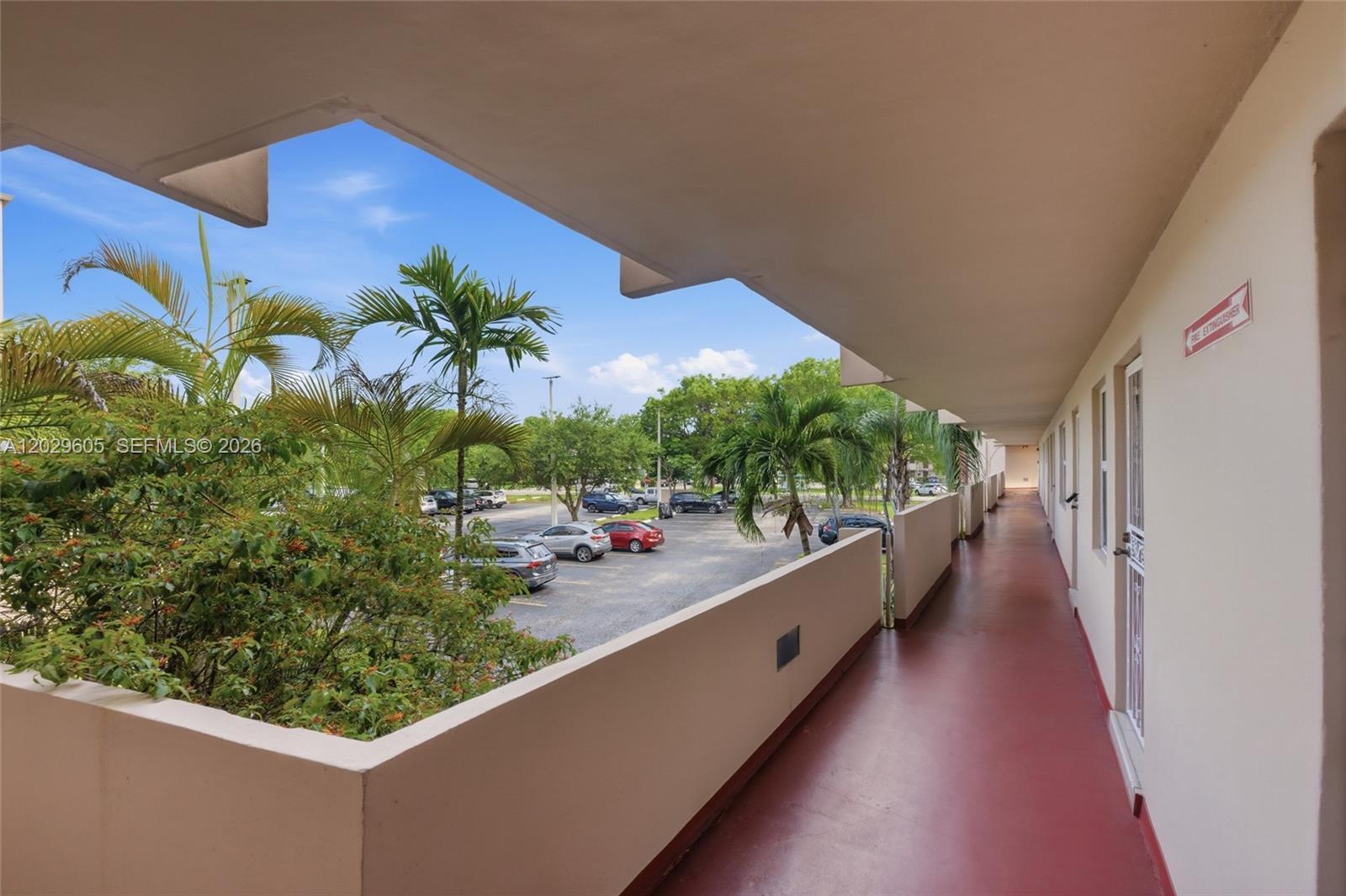 An outdoor apartment building walkway overlooks a parking lot with palm trees and cars — 1501 NE 191st St # C217, Miami FL 33179, MIAMI, FL